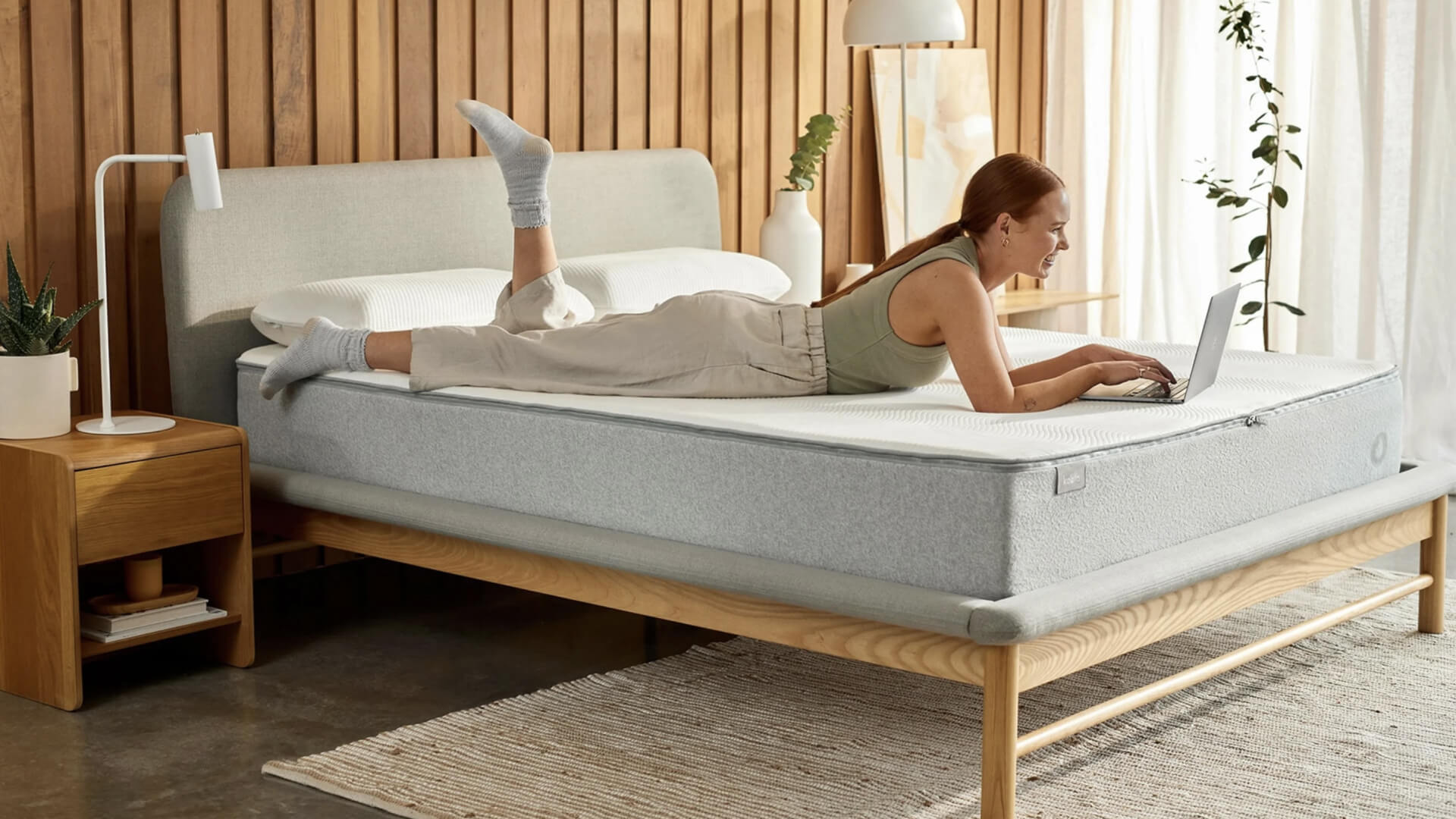 A woman lies on a light grey mattress atop a wooden bed frame, using a laptop. There’s a wooden bedside table with a lamp. Light wood panel wall.