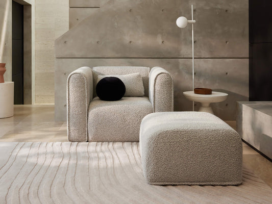 Modern lounge with a light grey armchair and matching ottoman, textured fabric, black and grey cushions, white floor lamp, and neutral rug.