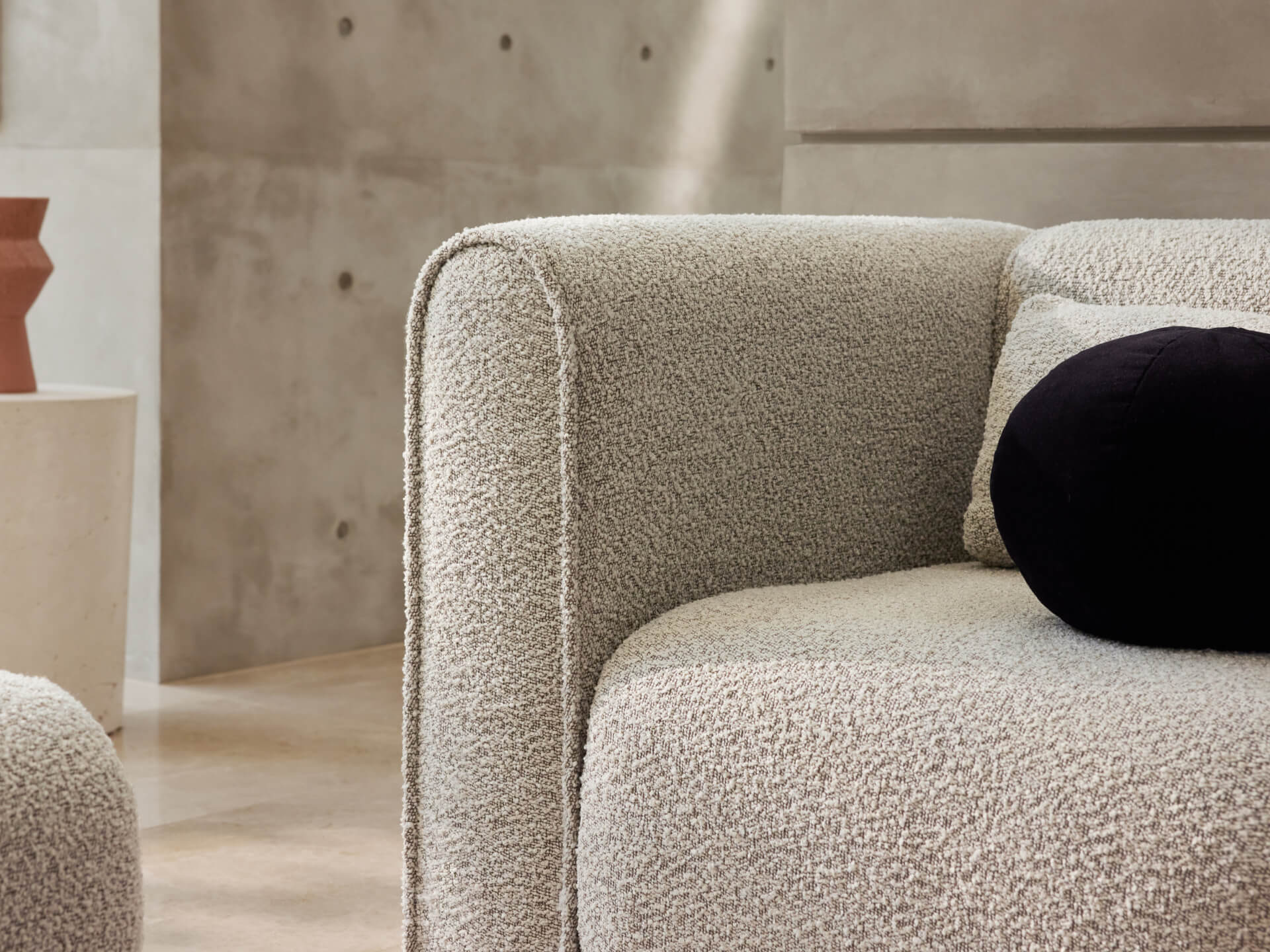 Close-up of a textured light grey couch with rounded edges, set against a concrete wall. A black cushion rests on the seat.