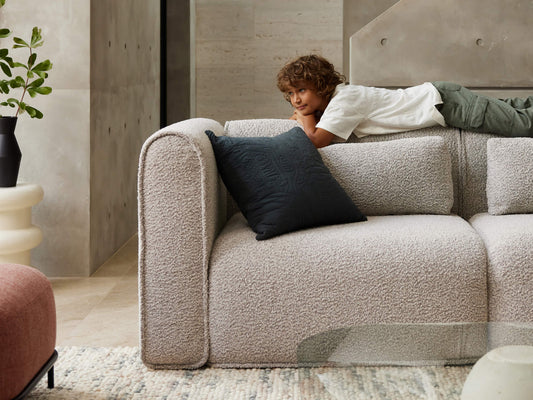 Boy lying on a light grey textured sofa with a black cushion in a modern living room. Beige walls, pink chair, and glass table nearby.