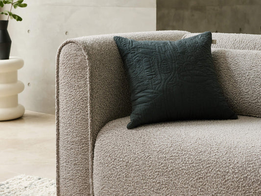 Light grey boucle sofa with a dark green textured cushion. Minimalist decor with a white sculptural side table and black vase in the background.