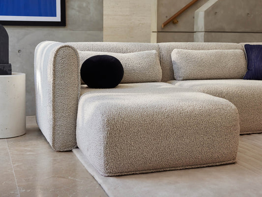 Modern light grey sectional sofa with textured fabric, featuring cushions and a black round pillow. Set on a tiled floor near a wall.