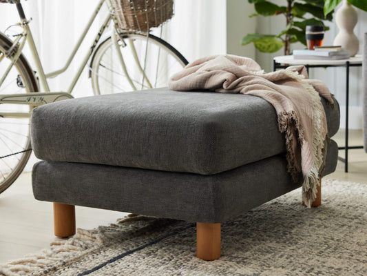 Dark grey ottoman with wooden legs, draped with a pink throw. Near a bicycle and a small table with plants, all on a textured rug.