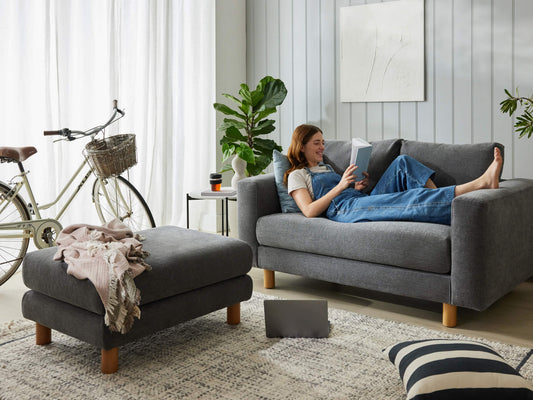 Woman relaxing on a grey sofa reading, with a matching ottoman nearby. Indoor plant, bicycle, and art enhance the cosy living space.