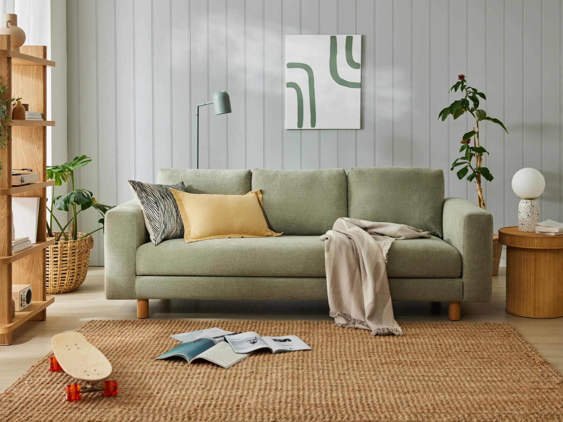 Sage green sofa with yellow and grey cushions, beige throw, and wooden legs. Features a jute rug, skateboard, side table, and potted plant.