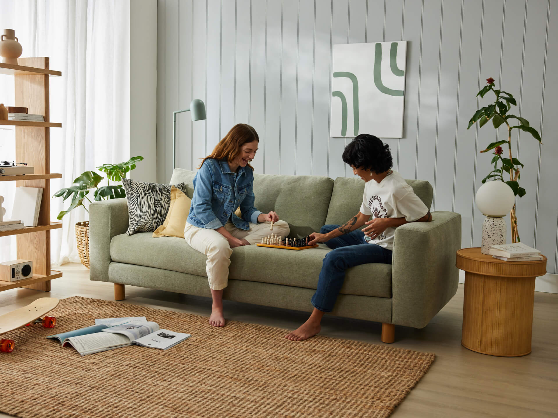Two people sit on a light grey sofa playing chess in a stylish living room with wooden shelves, greenery, and modern art.