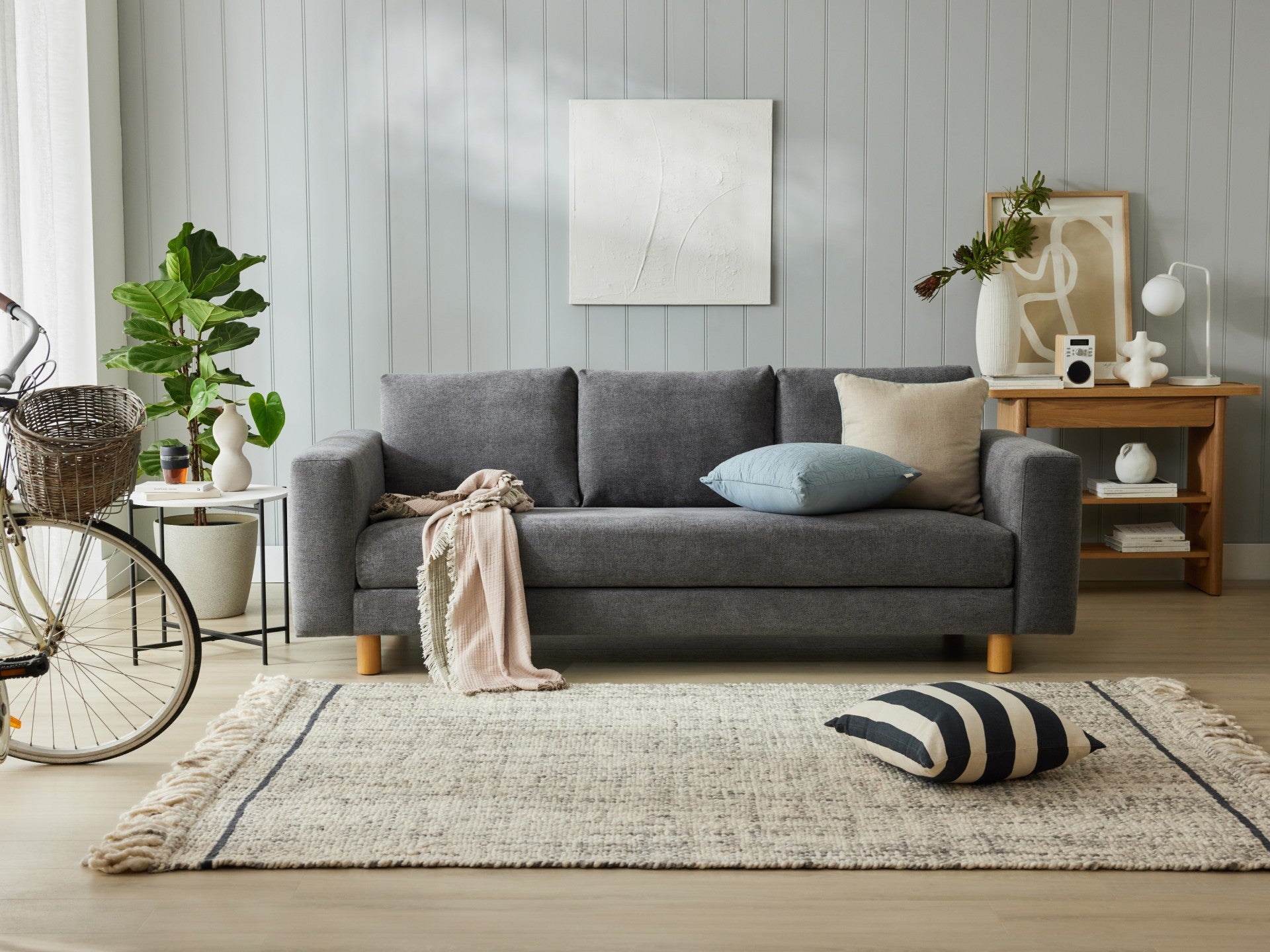 Modern living room with grey sofa, blue and beige cushions, and light grey rug. Wooden side table with decor; indoor plants and bike visible.