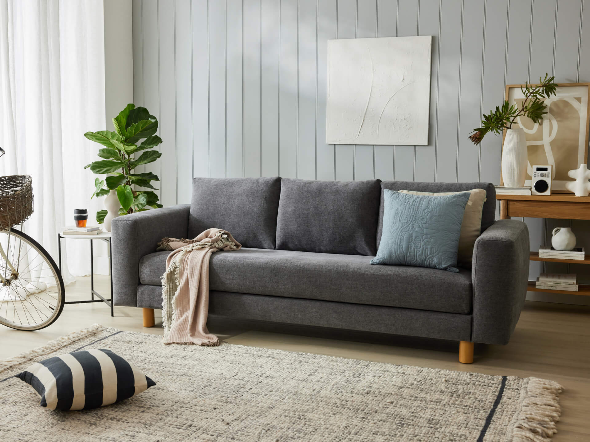 Modern living room with a grey couch, blue and beige cushions, and a throw blanket. Wooden accents, potted plant, and decorative items on a console.