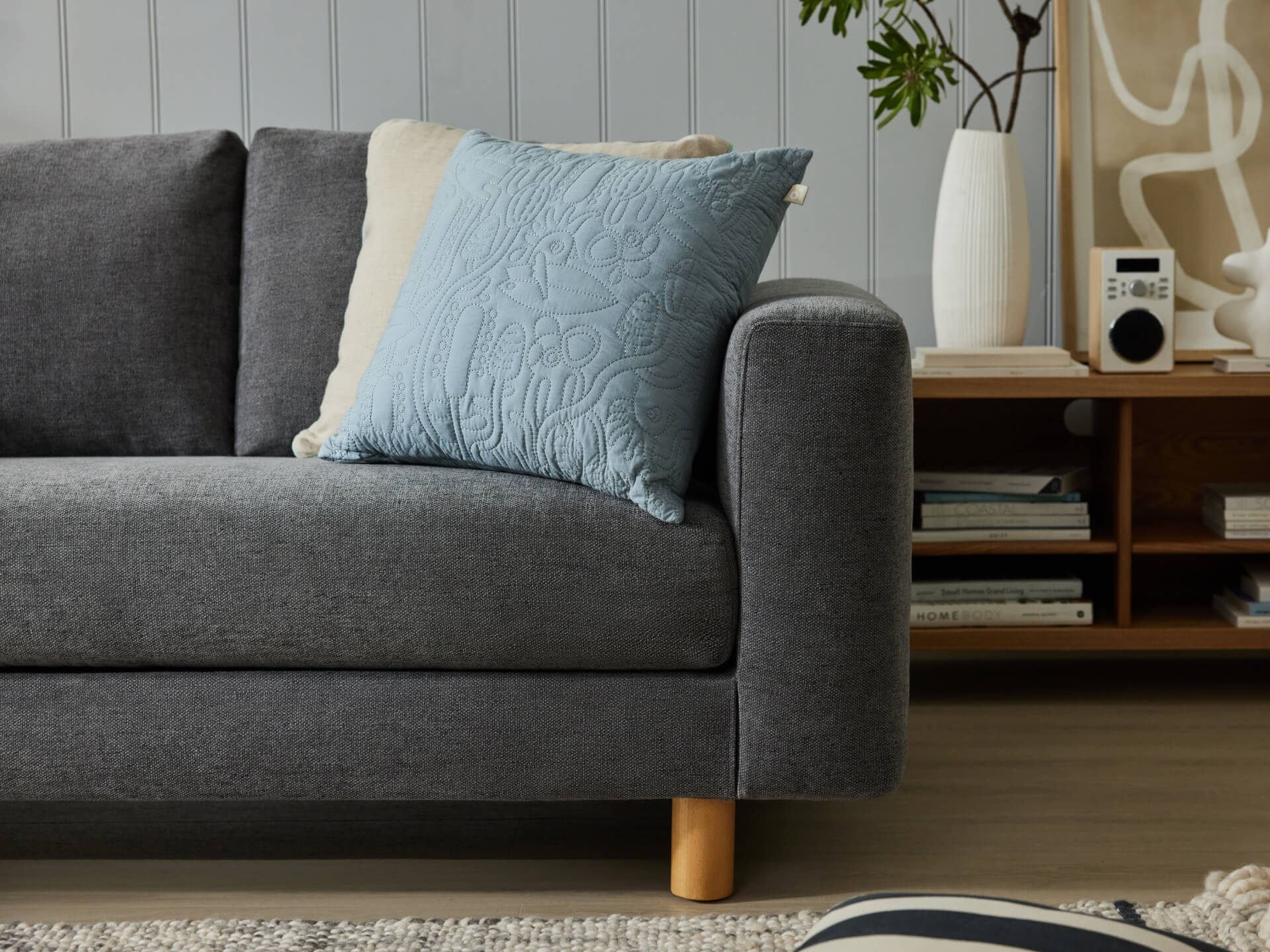 Dark grey sofa with light blue patterned cushion, light wooden legs. Wooden shelf behind with books, white vase, and radio. Rug on floor.