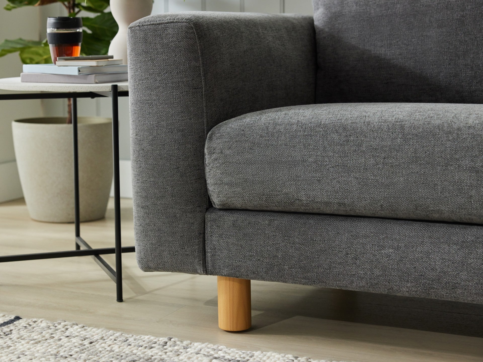 Close-up of a dark grey fabric sofa with light wood legs. Next to it, a modern side table with books, a plant, and a coffee cup on a light wood floor.