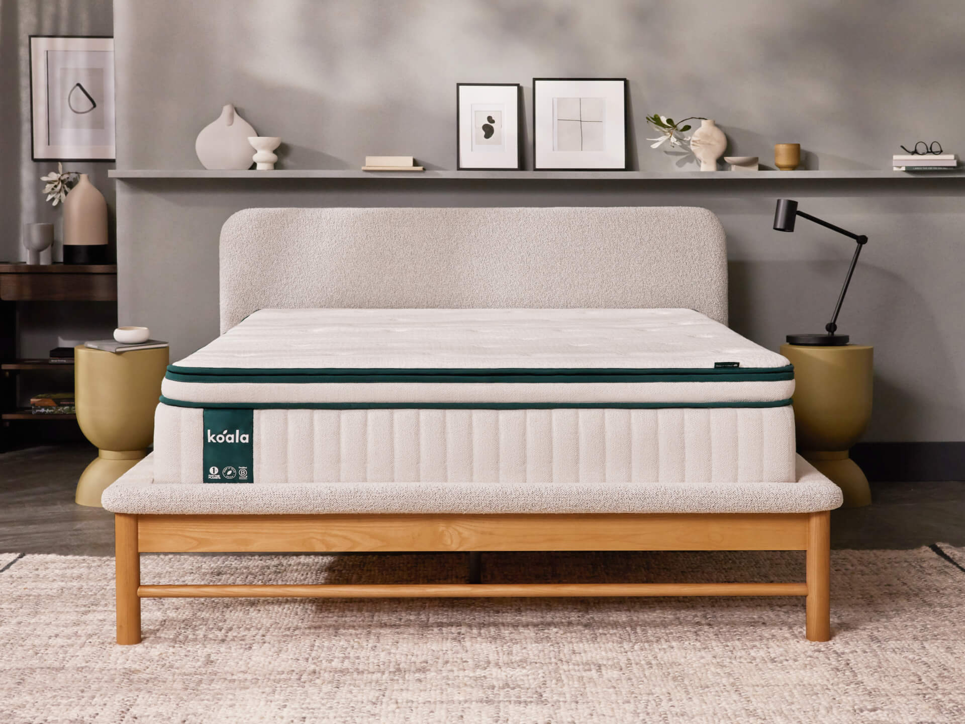 Beige bed with a white mattress and green trim in a modern bedroom. Neutral decor includes a shelf with art, vases, and a black lamp.