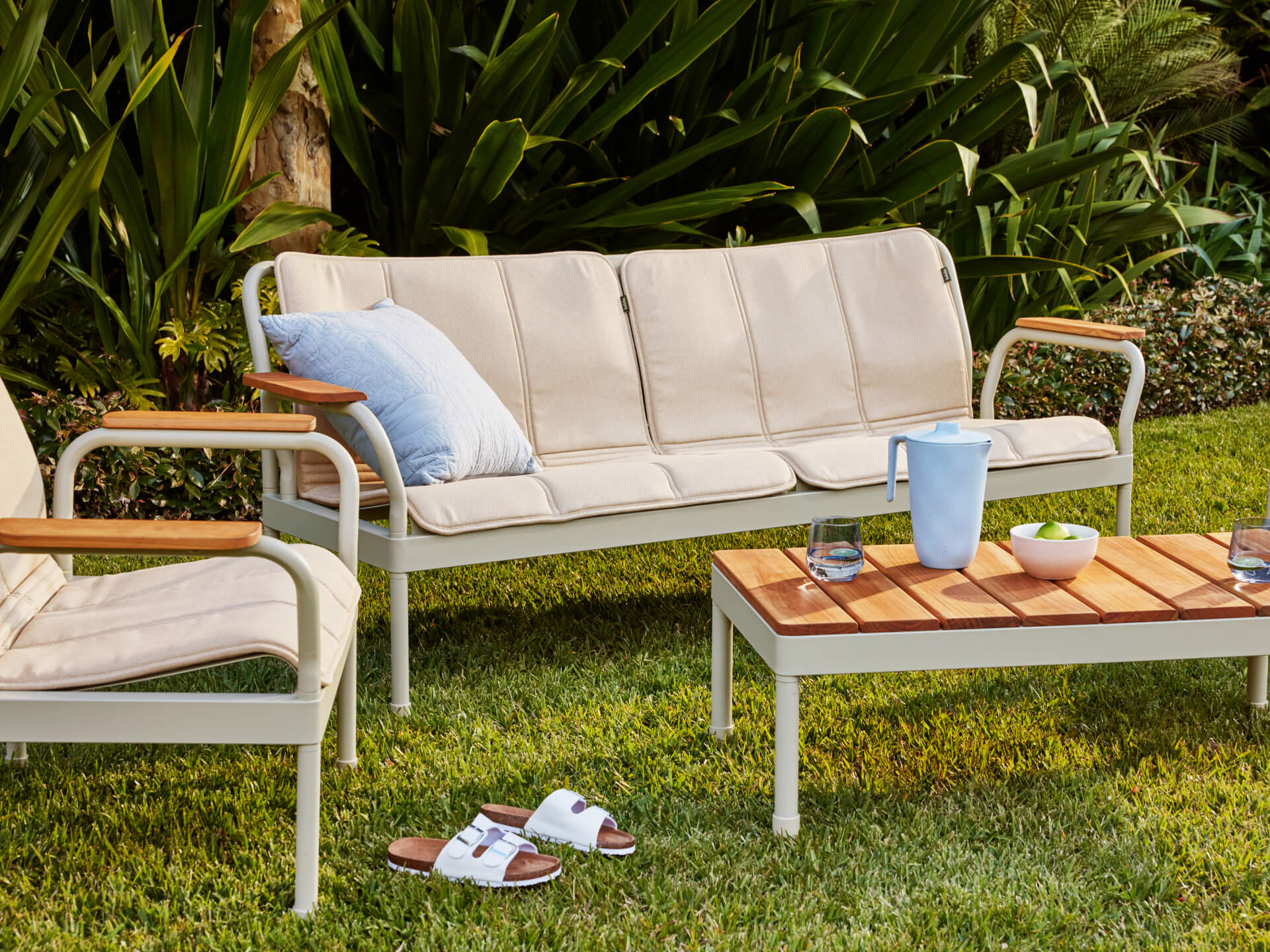 Outdoor setting featuring a cream three-seater couch with a blue cushion, matching chair, wooden table, and sandals on green grass.