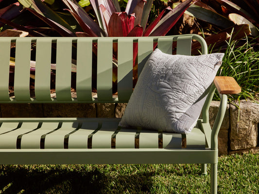 Mint green metal outdoor bench with wooden armrest, adorned with a light grey textured cushion. Surrounded by vibrant garden plants.