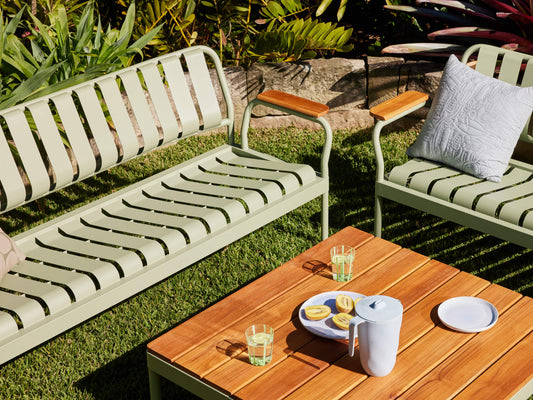 Outdoor setting with two light green metal benches on grass, a wooden table with drinks, lemons, a jug, and a cushion. Surrounded by plants.