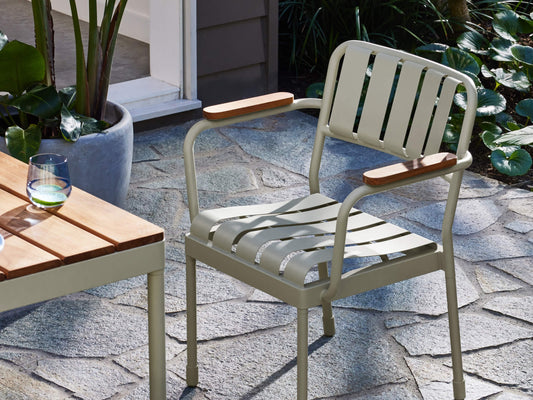 Outdoor setting with a light grey metal chair featuring wooden armrests on a stone patio. Nearby is a wooden-top table with a glass of water.