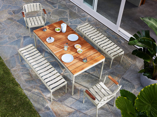 Outdoor dining set with a wooden table, two light grey slat benches, and two matching chairs. Plates and cups are set on a stone patio.