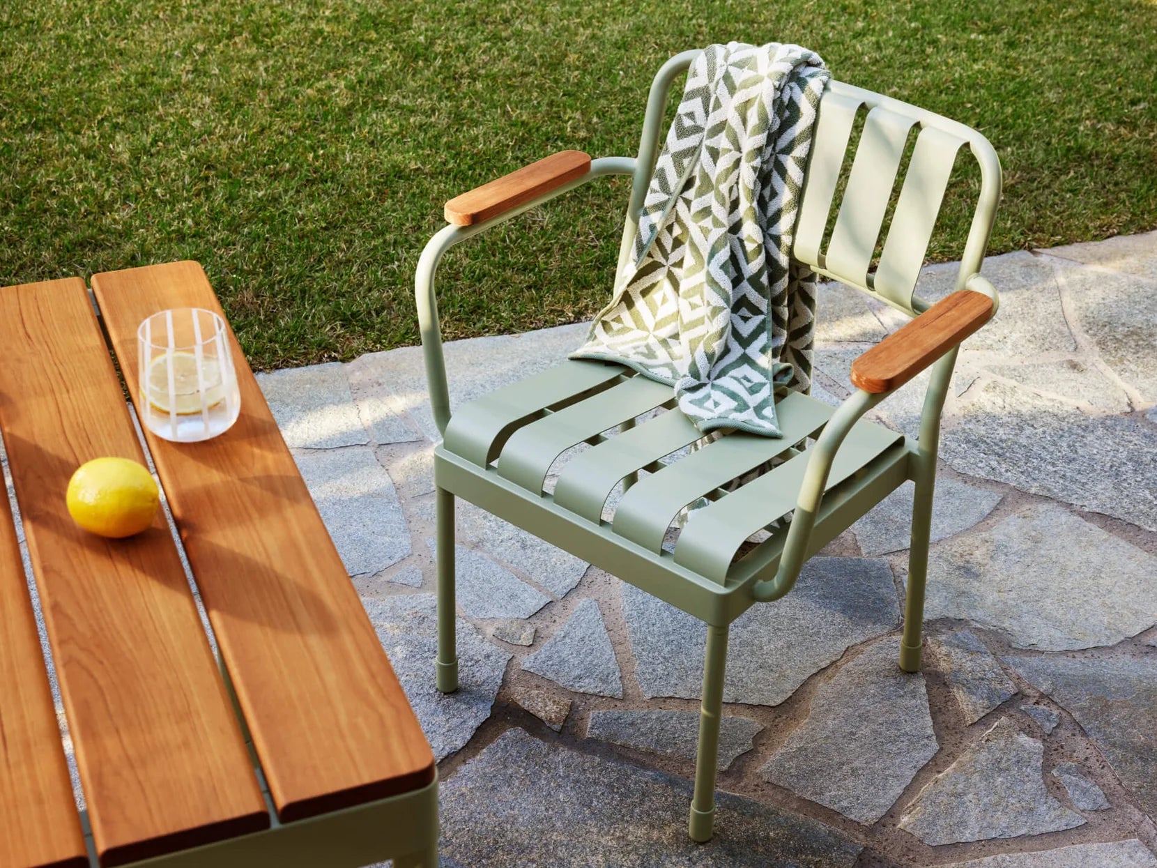 Green patio chair with wooden armrests on stone pavement, draped with a patterned blanket. Nearby, a wooden table holds a lemon and glass.