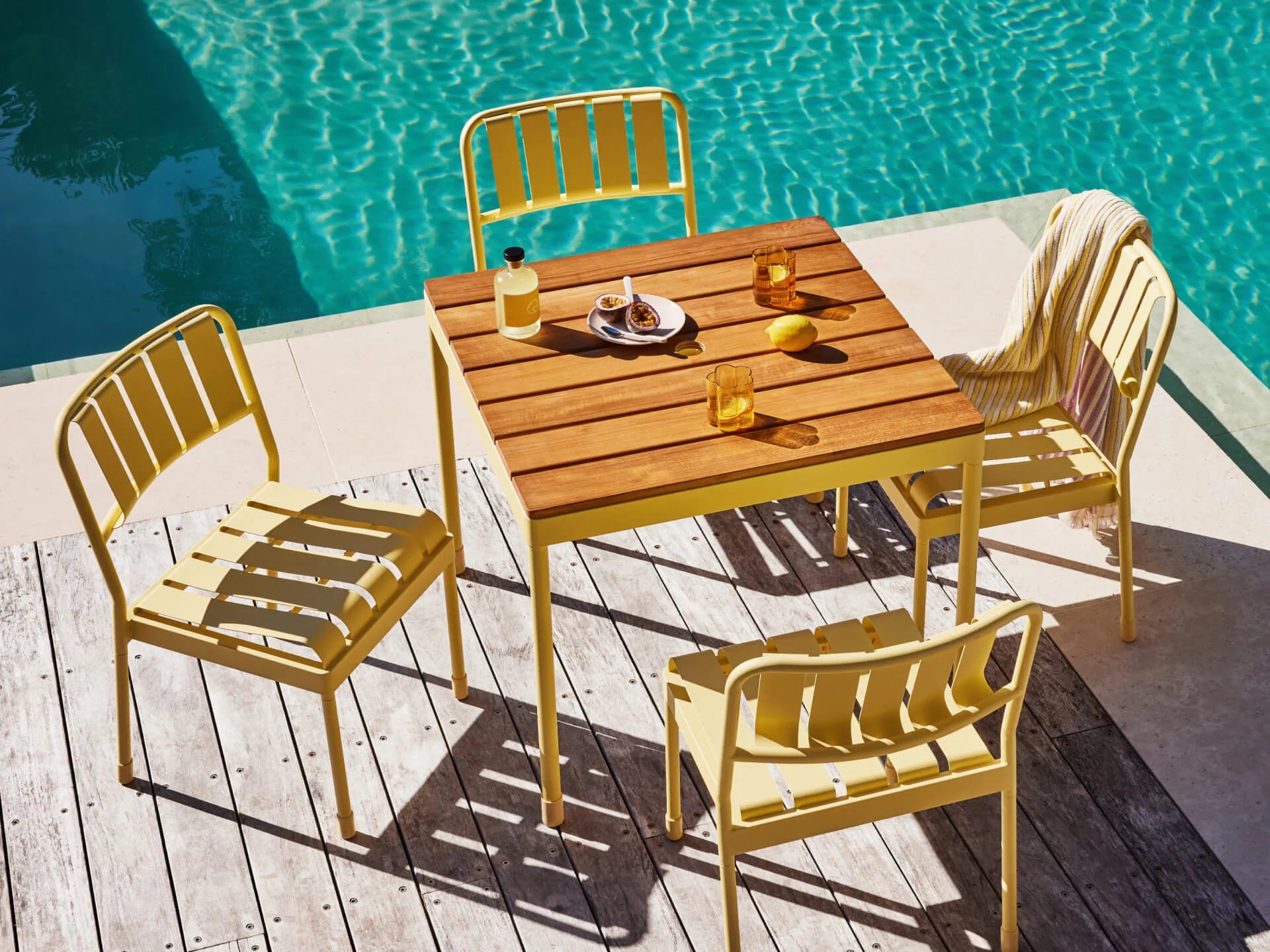 Yellow outdoor dining set with four chairs and a wooden table on a deck by a bright blue pool. Drinks and a towel are on the chairs.