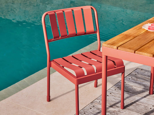 Coral chair with slatted design next to a wooden table, placed by a pool on light stone tiles.