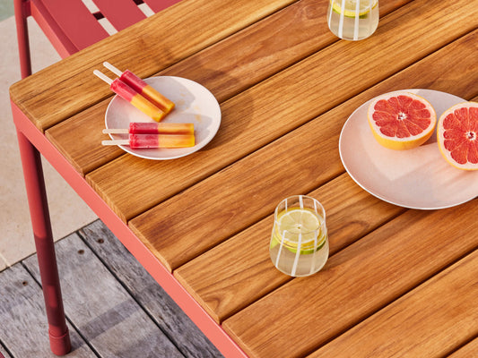 Wooden outdoor table with a red frame, holding a plate of ice blocks, halved grapefruit, and a glass of water, on a deck.