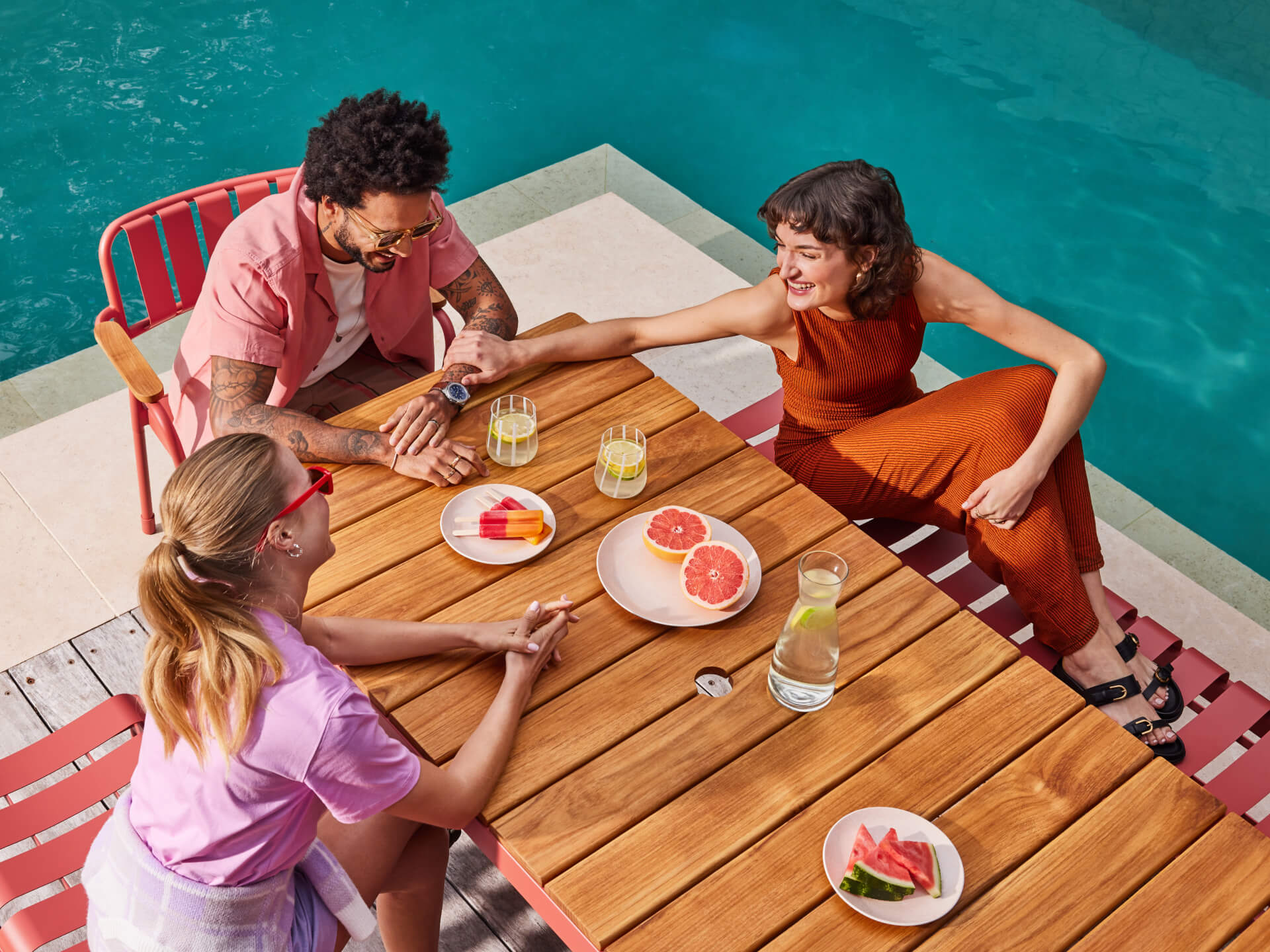 Three people sitting at a wooden outdoor table next to a pool, enjoying fruit and drinks. Red chairs, casual atmosphere, sunny day.