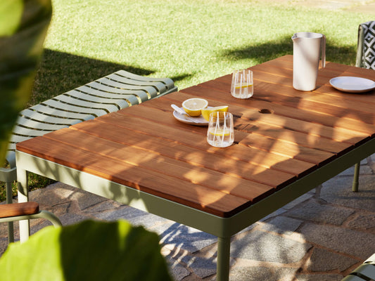 Wooden outdoor table with a green metal frame on a stone patio. Set with a jug, glasses, and a plate of lemons. Green grass and plants nearby.