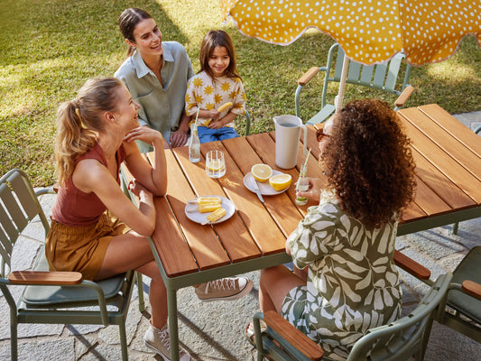 Four people enjoy drinks and snacks around a wooden outdoor table with green chairs. A yellow polka dot umbrella shades the setting.