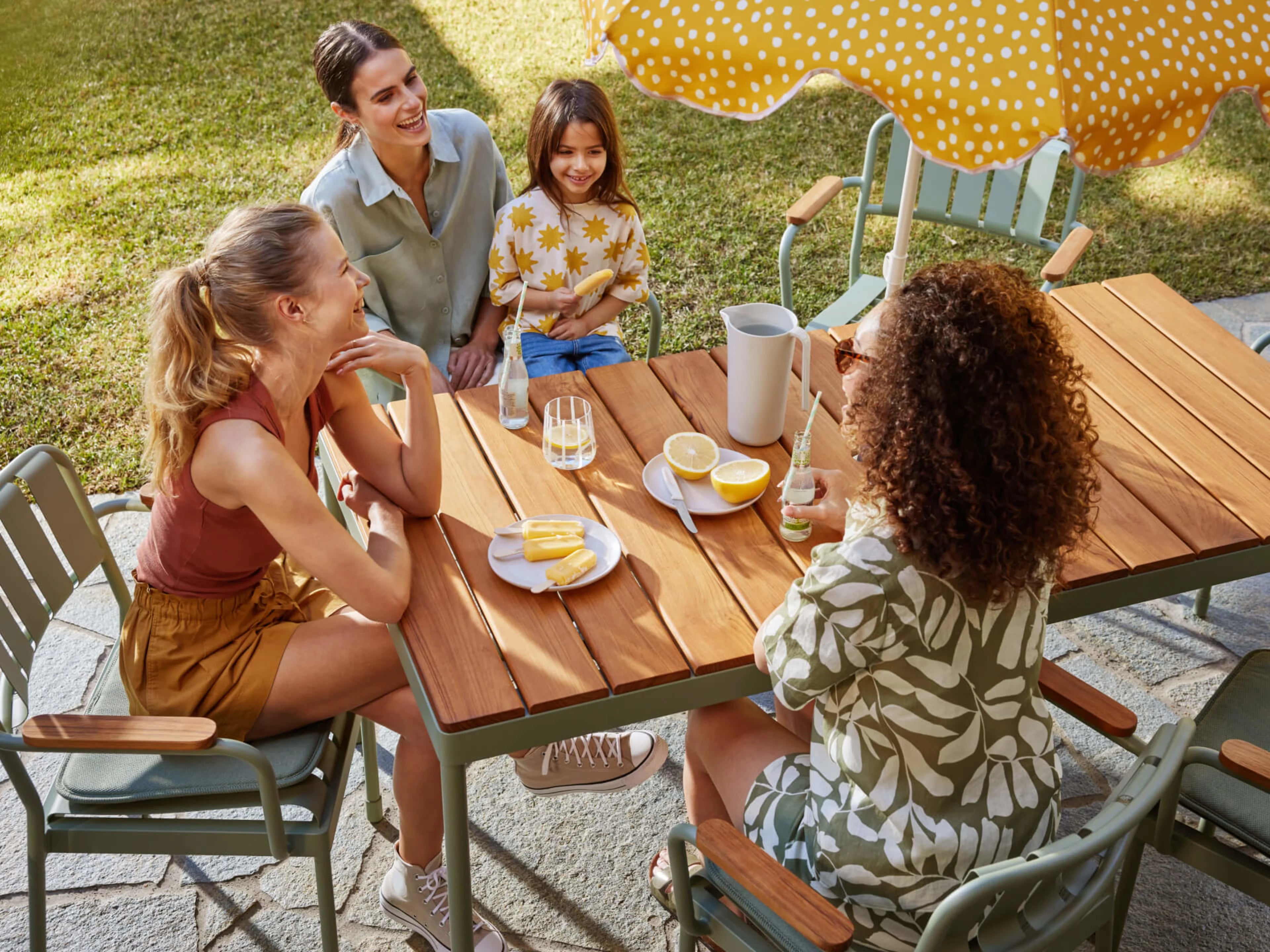 Four people enjoy refreshments at a wooden outdoor table with green chairs, under a yellow polka dot umbrella on a sunny day.