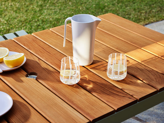 Wooden outdoor table with a white jug and two glasses of lemon water. Sliced lemons on a white plate. Grass in the background.