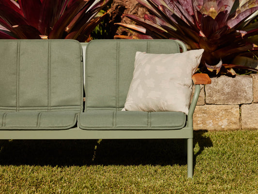 Outdoor green two-seater sofa with a light grey cushion, set on grass. Stone wall and lush plants in the background.