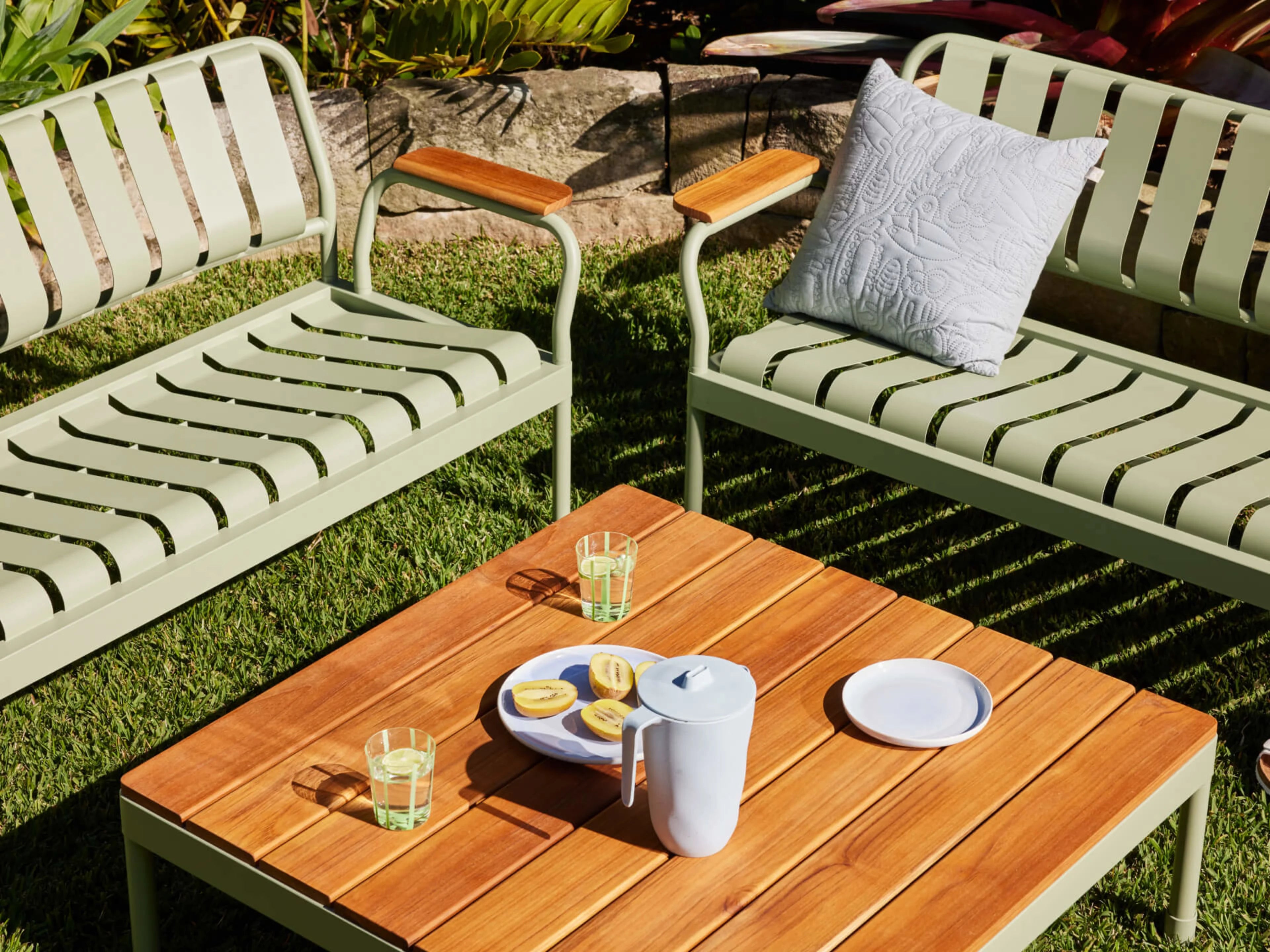 Outdoor setting with two light green metal benches and wooden armrests. A wooden table sits between them holding glasses, a jug, and a plate with lemons.