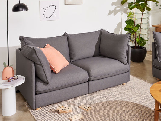 A dark grey sofa with plush cushions and a peach pillow on a light rug, surrounded by a potted plant, wall art, and a small side table.