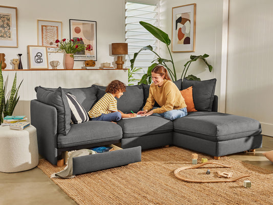 Living room with a dark grey L-shaped sofa featuring storage drawers, decorated with plants, framed art, and a woven rug on the floor.