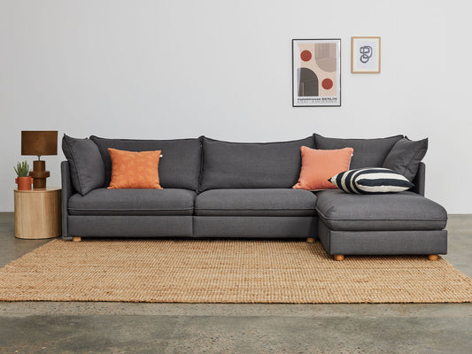 Dark grey sectional sofa with orange and striped cushions on a woven rug. Beside it, a wooden side table with a lamp and plant. Minimalist art on the wall.