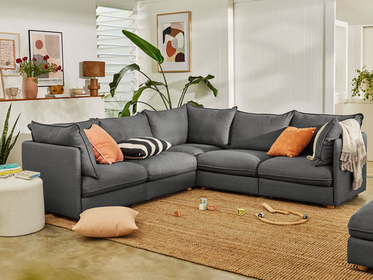 A dark grey sectional sofa with colourful cushions on a textured rug. Nearby, indoor plants, wall art, a side table, and toys embellish the space.