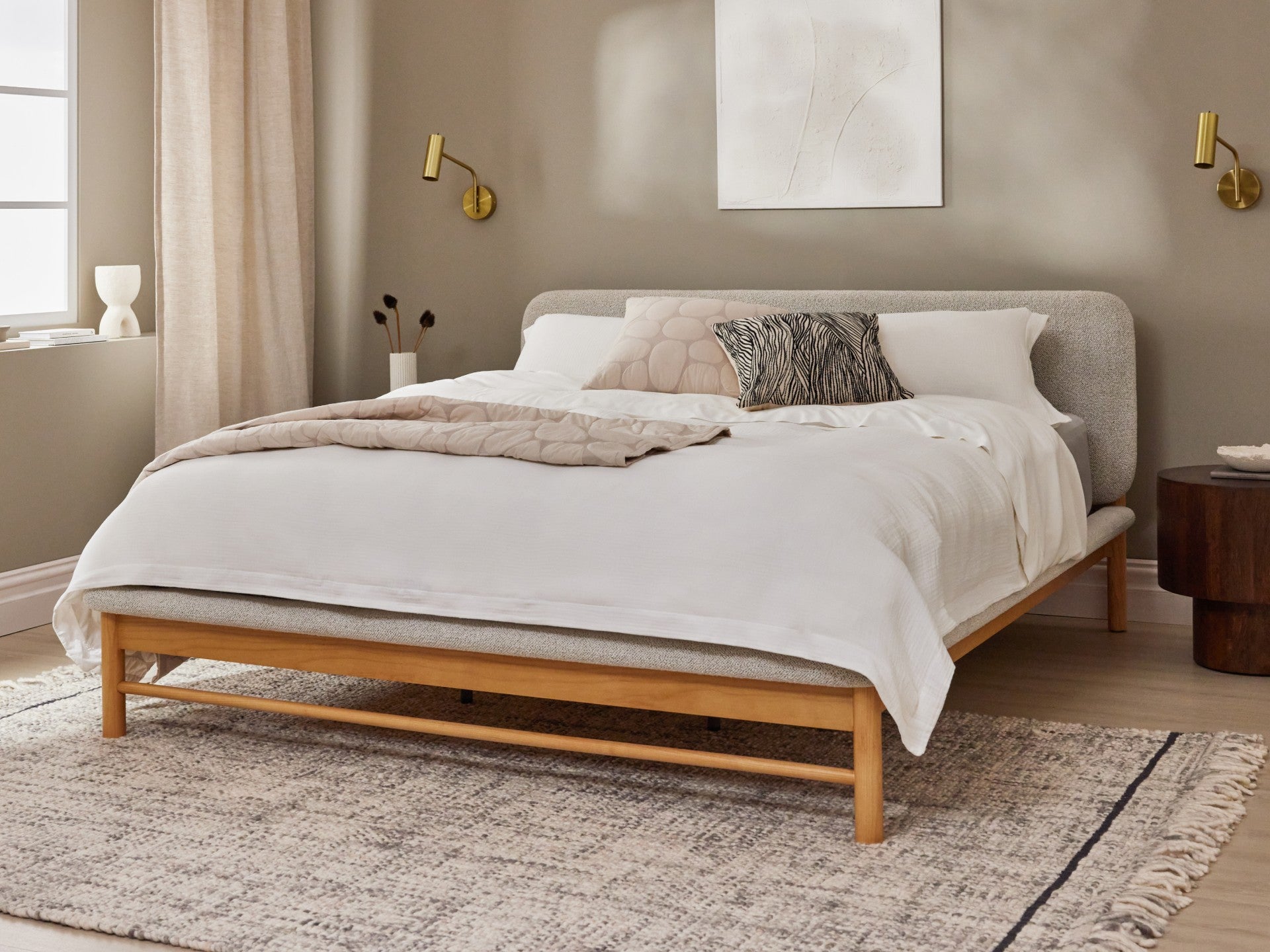 Minimalist bedroom with a wooden bed frame, white bedding, grey headboard, and beige walls. Soft lighting, neutral decor, and a textured rug.