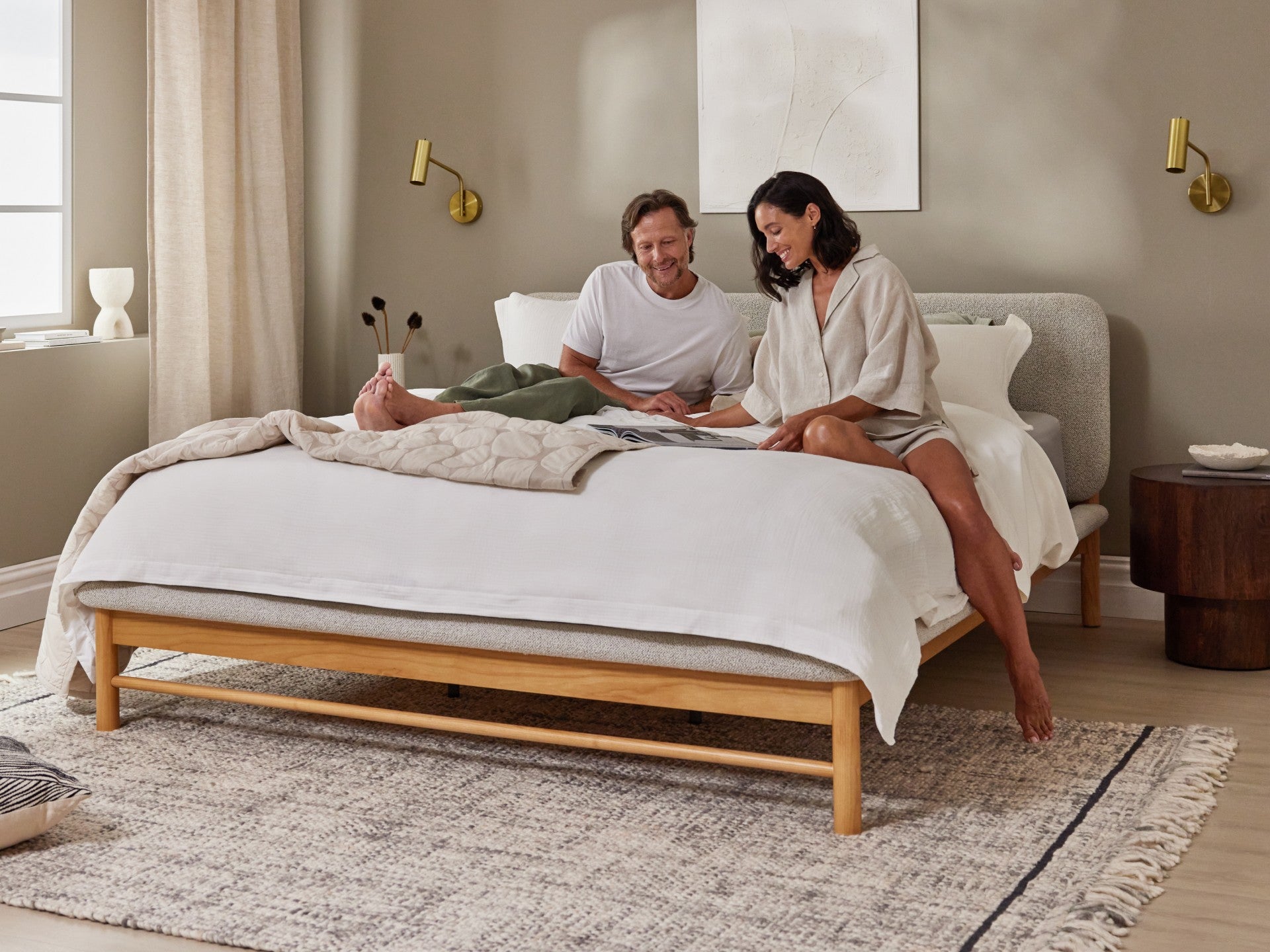 Couple sitting on light grey upholstered bed with wooden frame, white bedding, beside a round wooden side table and beige rug in a cosy bedroom.