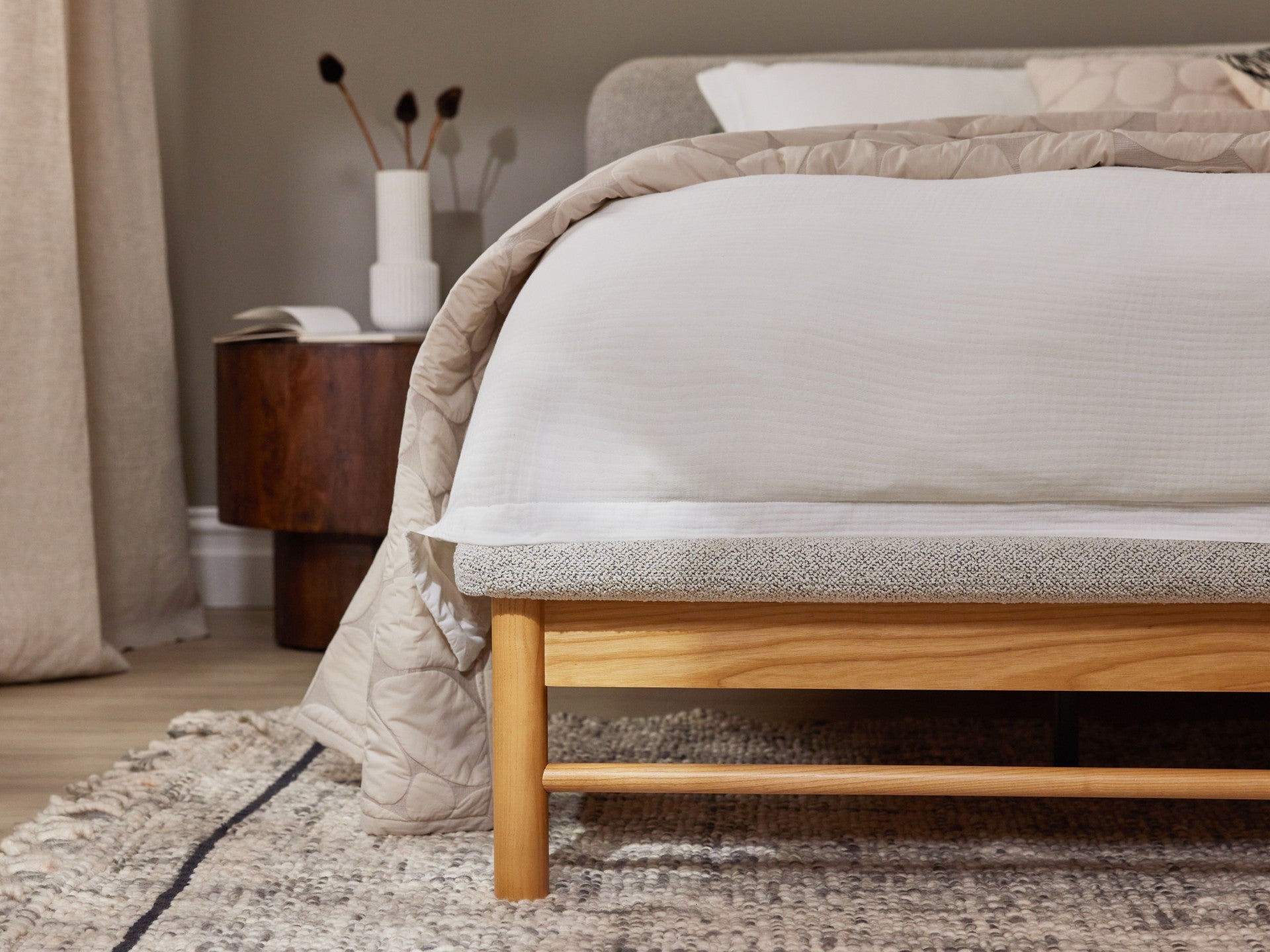 Bedroom with a wooden bed frame, white and beige bedding, a round wooden side table, and decor vase. Neutral tones and textured rug.