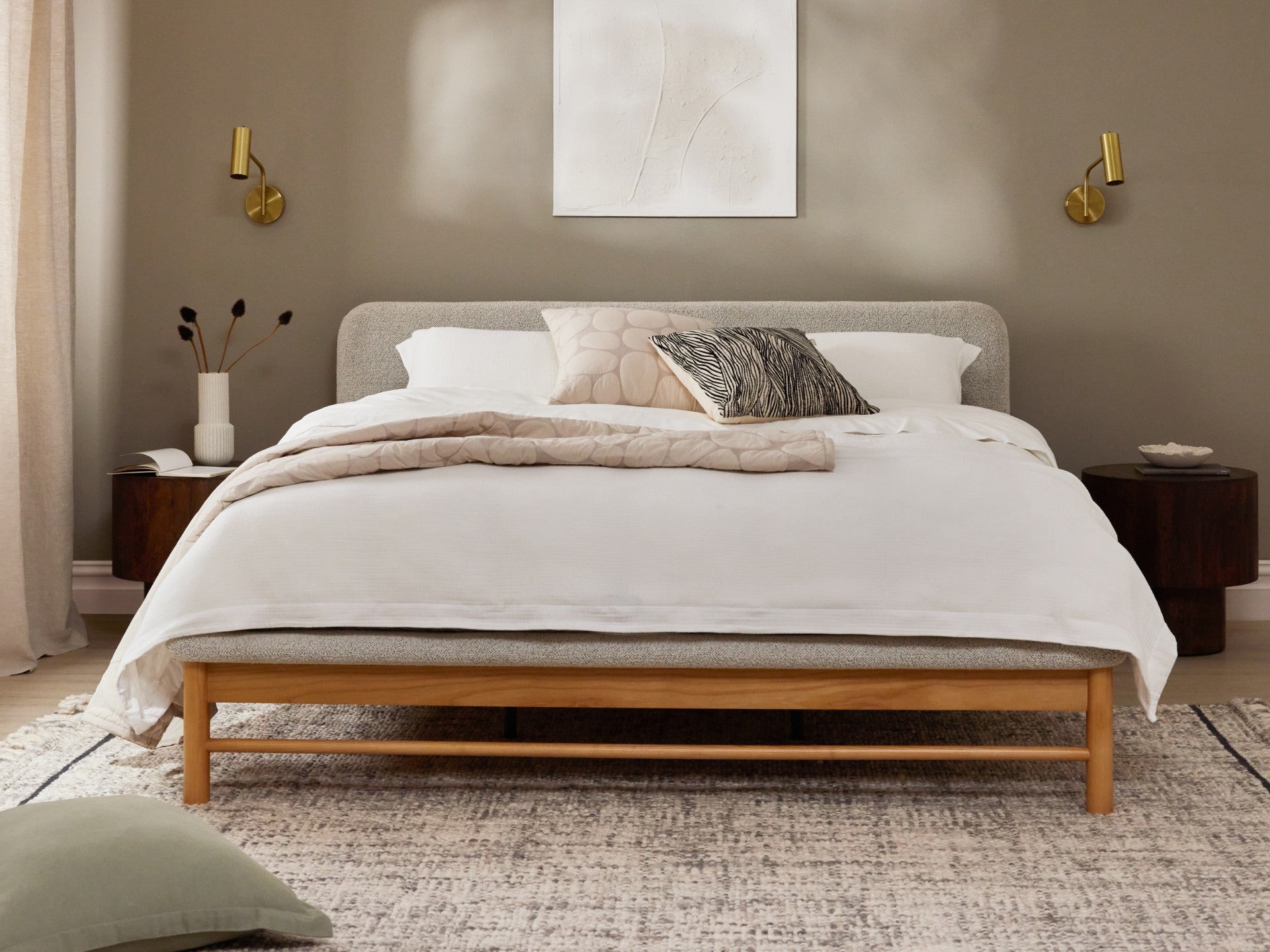 Minimalist bedroom with a wooden bed frame, neutral bedding, and textured cushions. Modern sconces and side tables complement the serene decor.