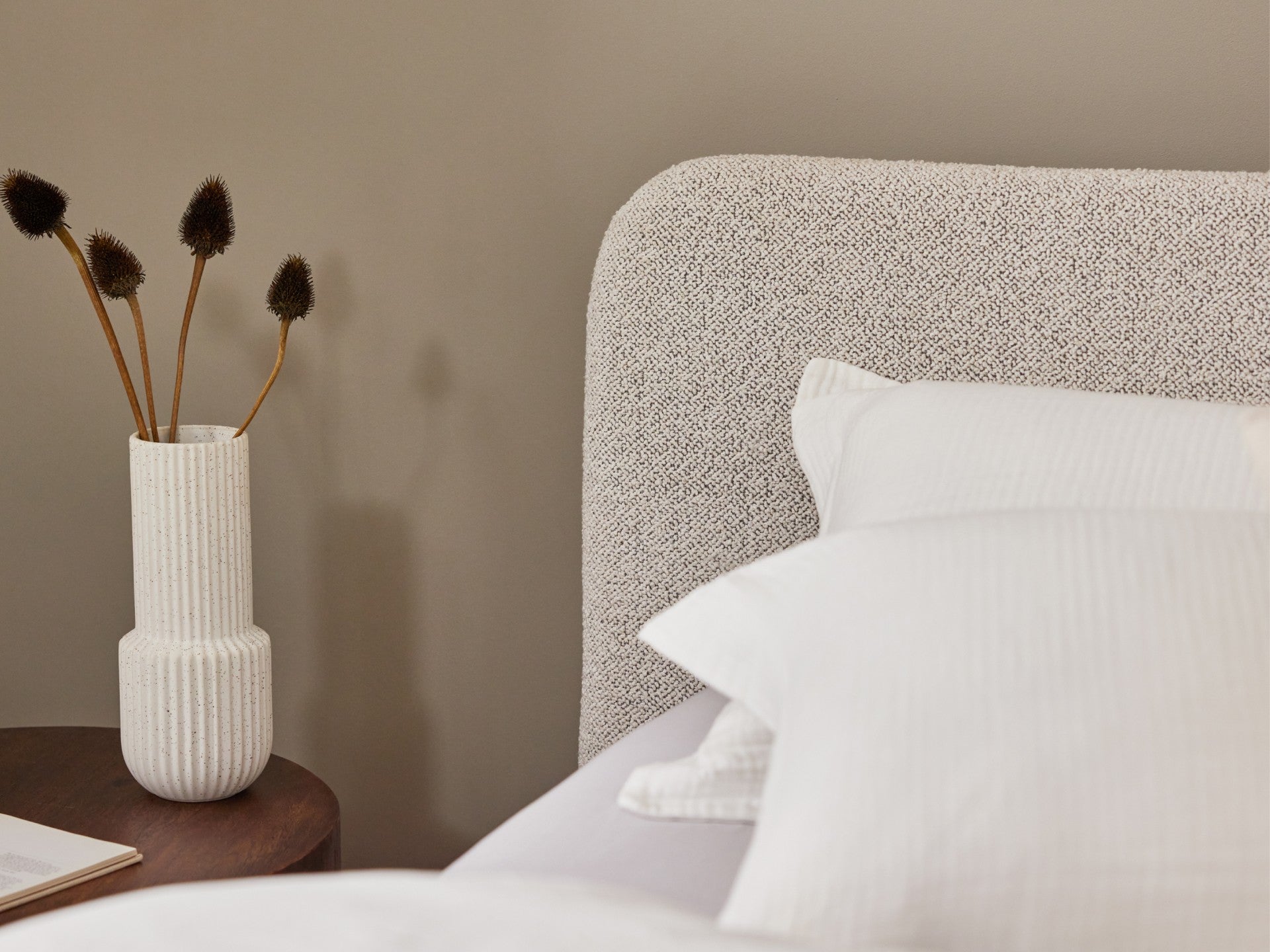 Beige upholstered bedhead with white pillows, next to a round wooden table featuring a textured white vase holding dried flowers.