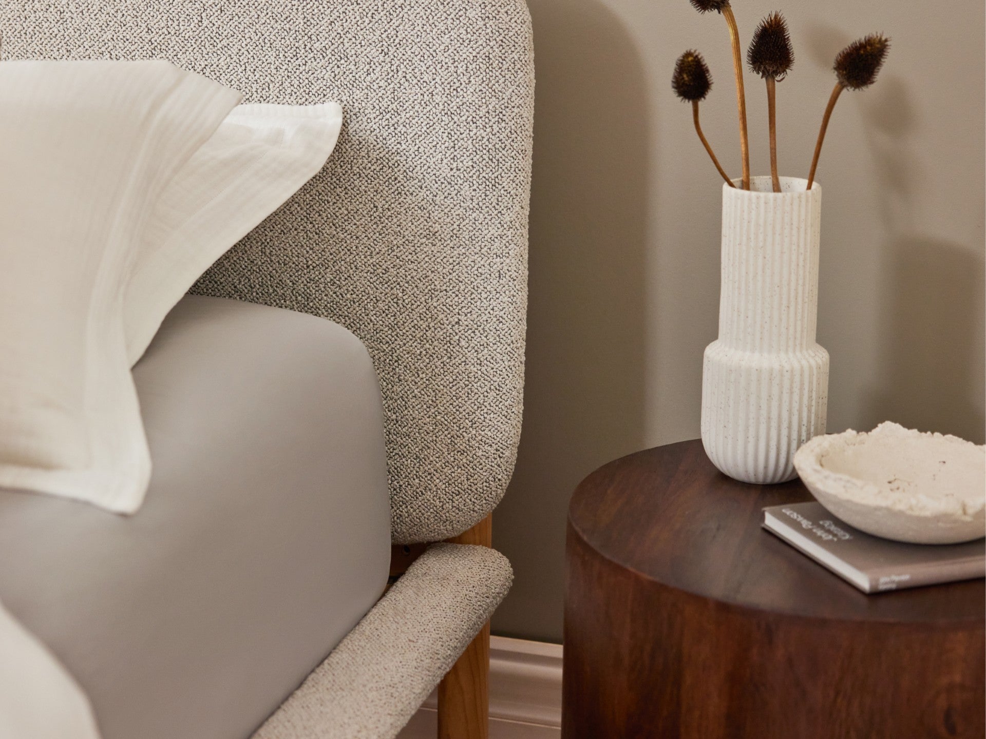 Bed with light grey headboard, grey sheet, white pillow, beside a dark wooden nightstand with a white vase of dried flowers and a decorative bowl.
