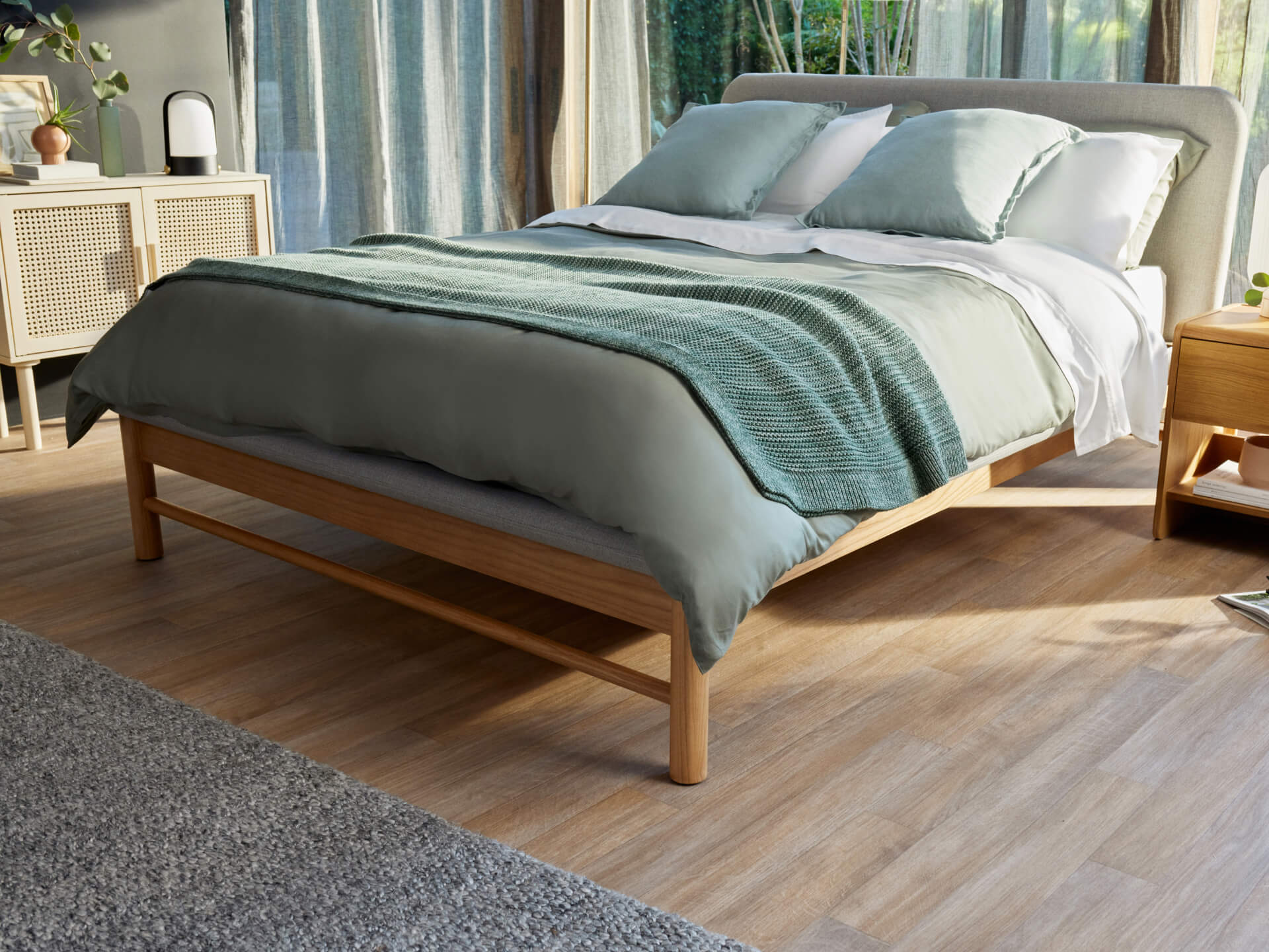 Modern bedroom with wooden bed frame, grey upholstered headboard, light green bedding, white and green cushions, wooden floor, grey rug.