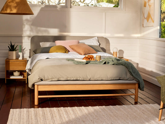 Cosy bedroom with a wooden bed, grey and green bedding, pink and yellow cushions, and wooden side tables on dark timber flooring.