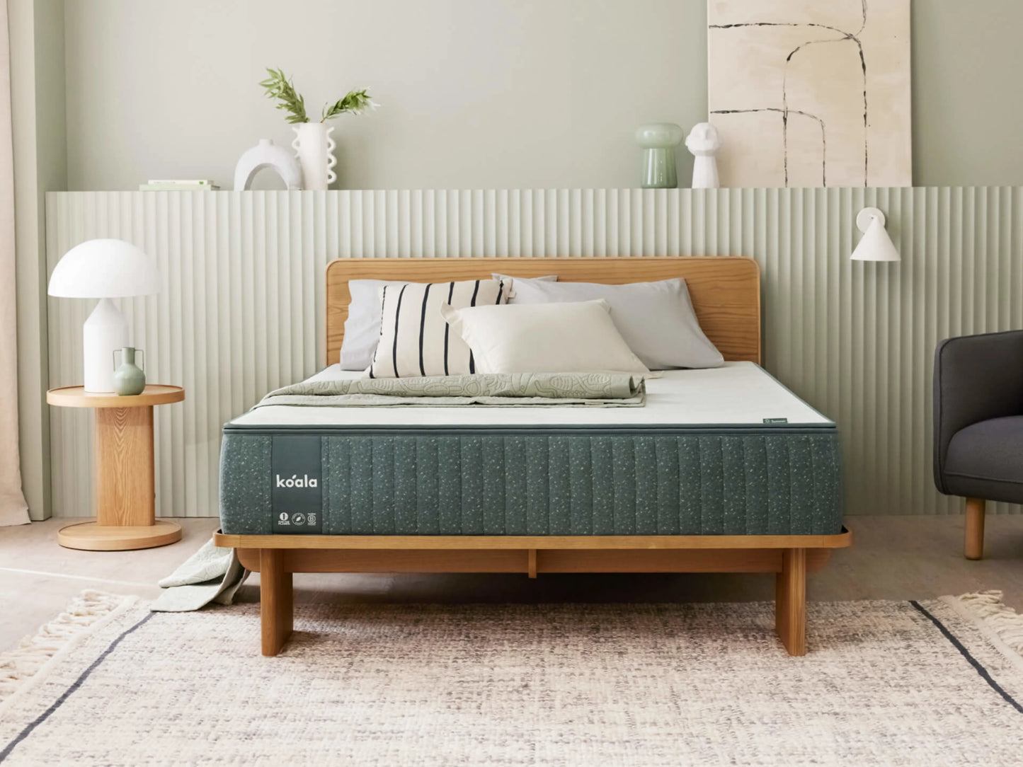 Modern bedroom with a wooden bed frame, green mattress, and assorted cushions. Light green wall, round side table, and decor objects adorn the room.