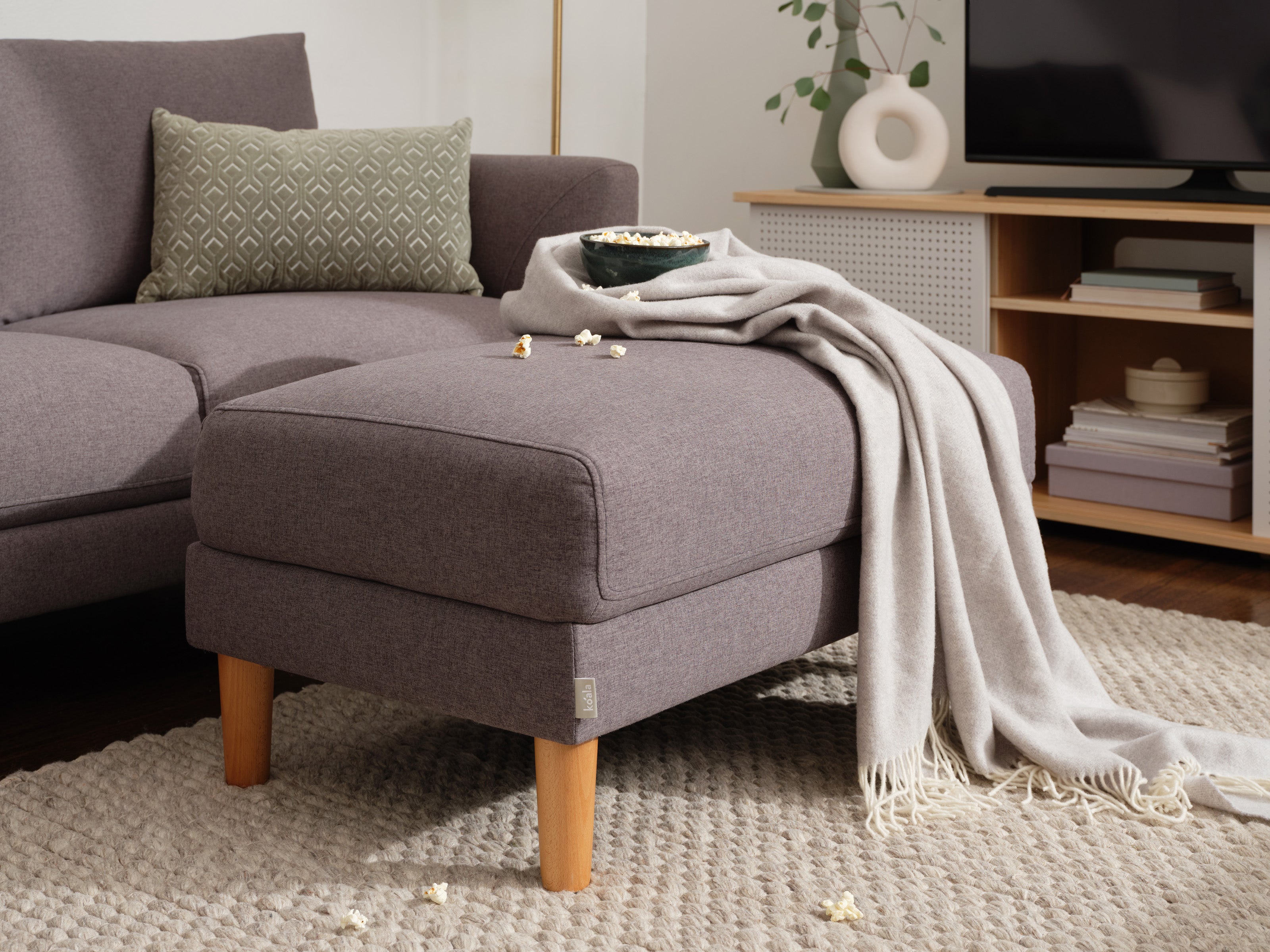 Grey sectional sofa with a matching ottoman, a green patterned cushion, beige throw, and popcorn bowl on top. Wooden legs and neutral decor.
