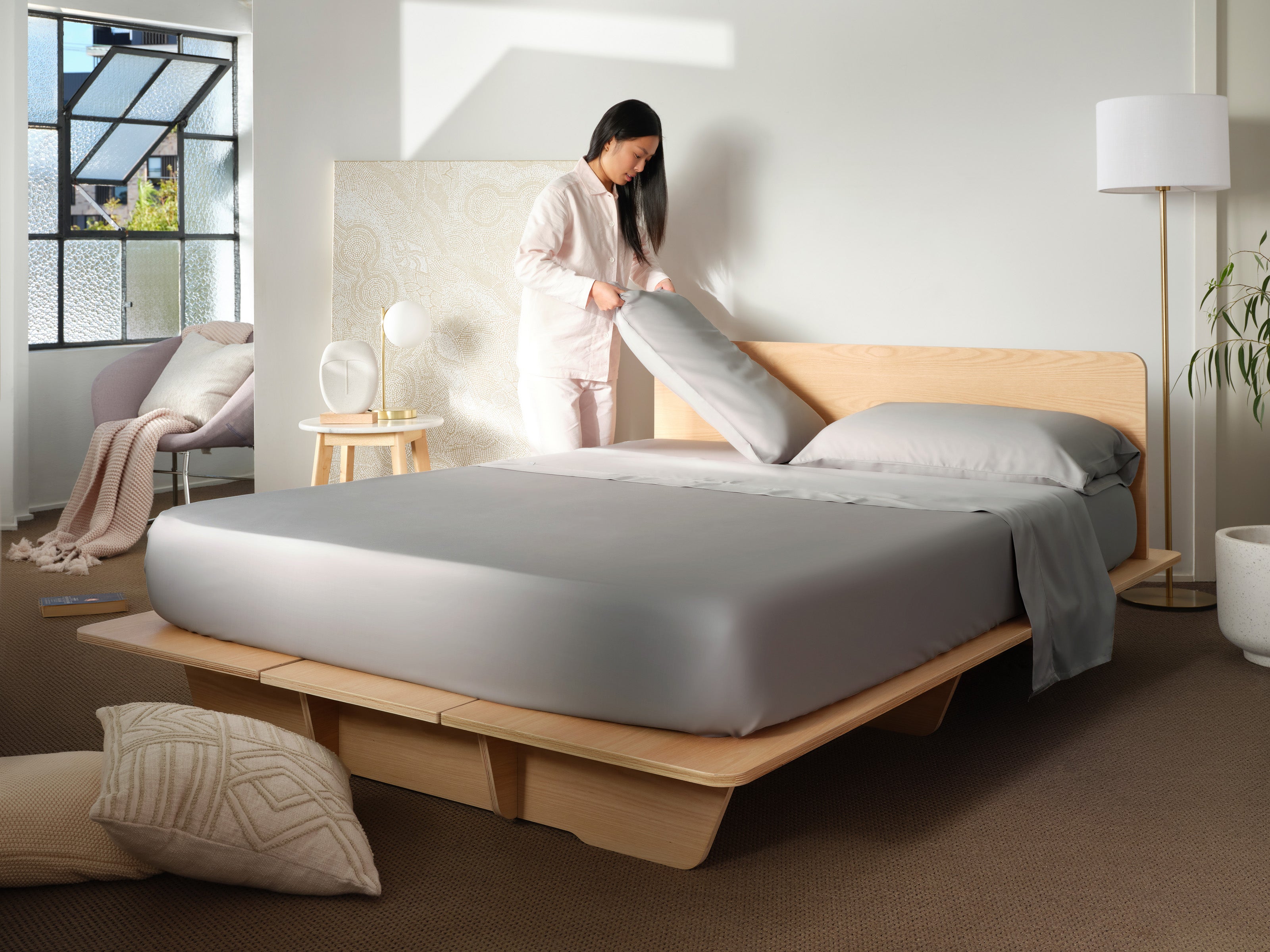 Modern bedroom with a light wood platform bed, grey bedding, and a person arranging pillows. Soft lighting, indoor plant, and a chair.