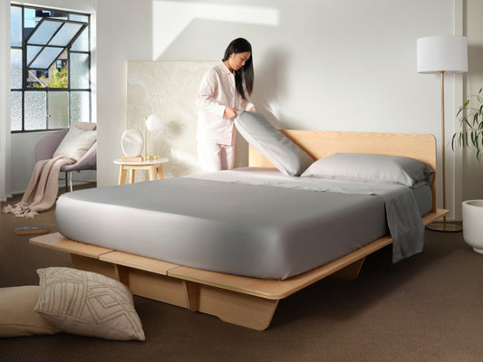 Modern bedroom with a light wood platform bed, grey bedding, and a person arranging pillows. Soft lighting, indoor plant, and a chair.