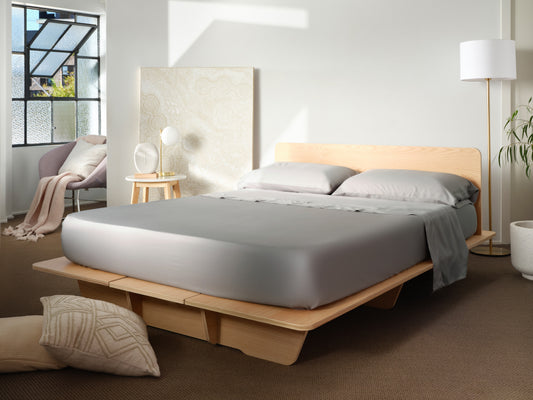 Minimalist bedroom with a light wood platform bed, grey bedding, and beige decor. A reading nook with a chair and large window enhances natural lighting.