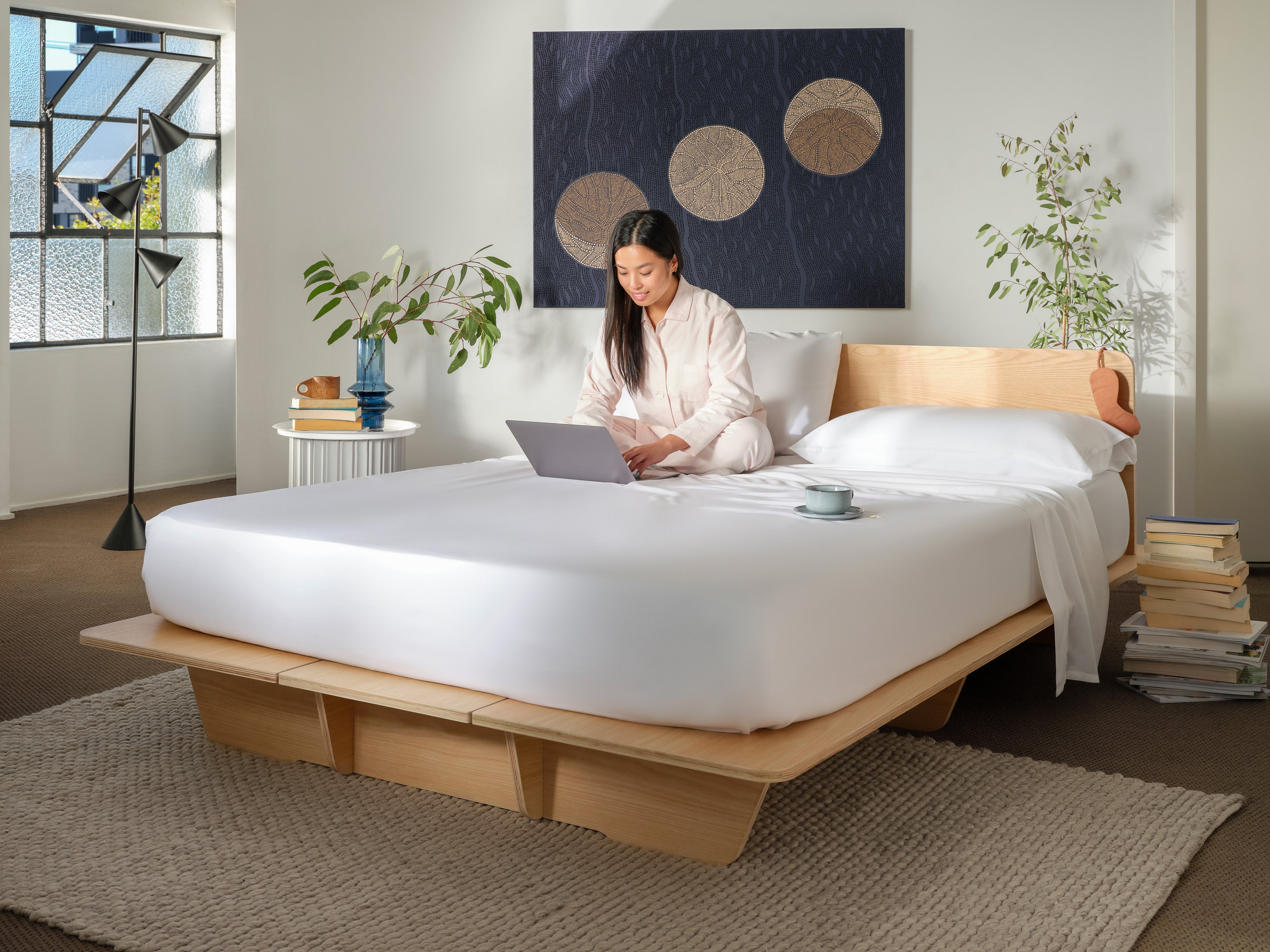 A woman uses a laptop on a light wood platform bed with white sheets in a modern bedroom. Plants, books, and a round art piece add decor.