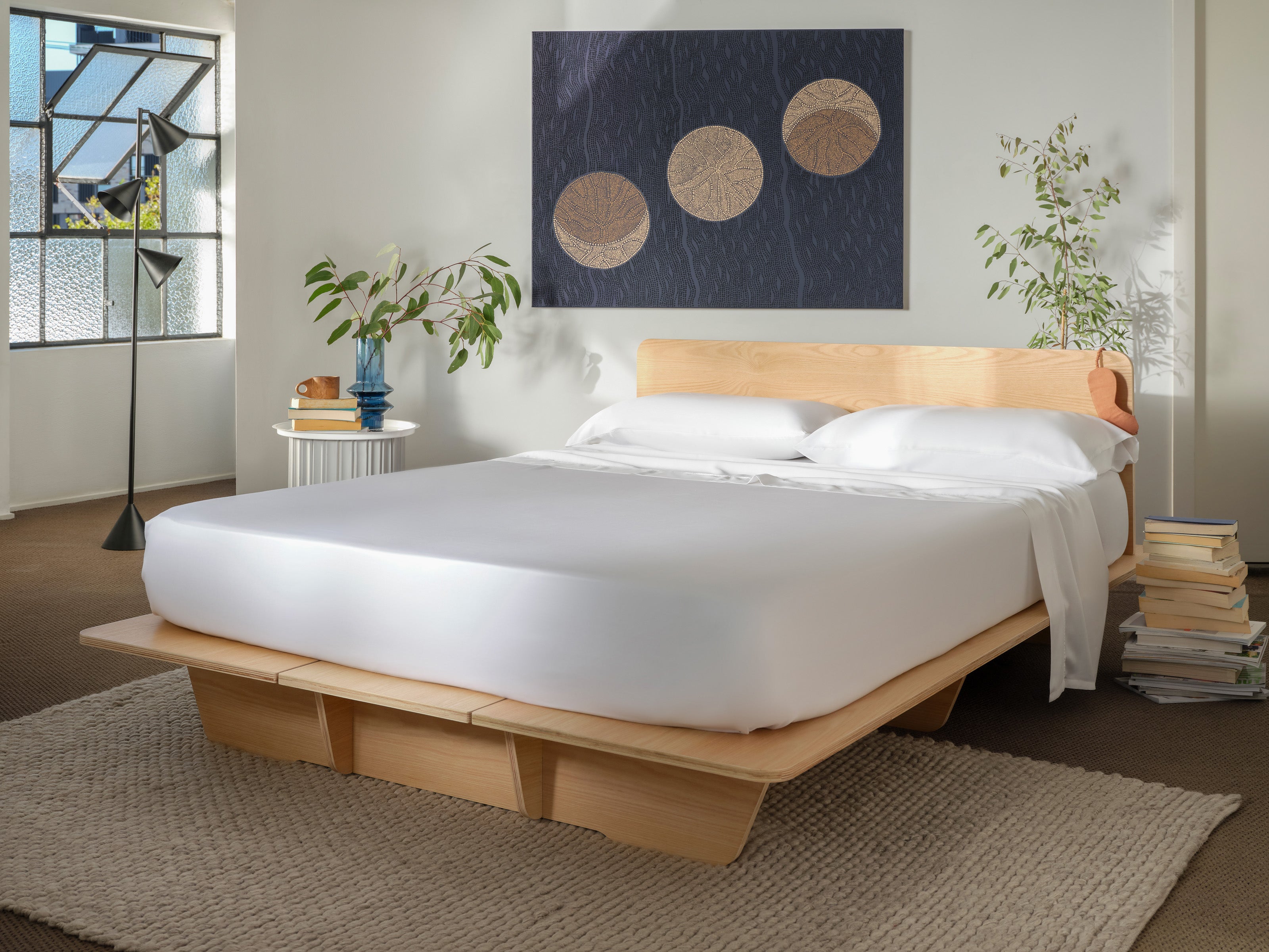 Minimalist bedroom with a light wooden platform bed, white bedding, abstract wall art, leafy plants, and a textured beige rug.